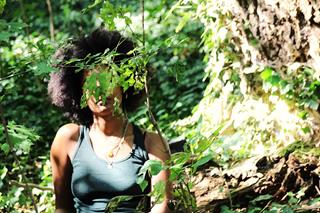 Summer forest with greenery and sunlight. A Black woman with afro hair is behind a tree branch, her face hidden by leaves.