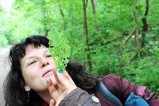 A woman with loose hair smiling closely at a green, lace-like leaf held by another person.