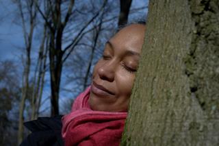 A dark-skinned woman in a forest, wearing a scarf, resting her head against a tree with closed eyes and smiling.