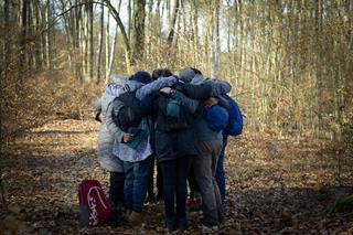In a winter forest on a hill surrounded by leafless trees, a group of people in warm clothing stands closely together in a circle.