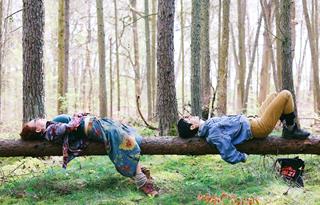 Winter forest in the sunlight, with bare trees and no undergrowth. Two women lie on a fallen tree, looking up at the sky.