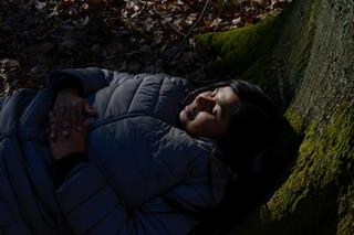 A woman from India with a torch, wearing a warm jacket, lying on the ground under a tree. Her face is in the sunlight, body in the shade, eyes closed, smiling.