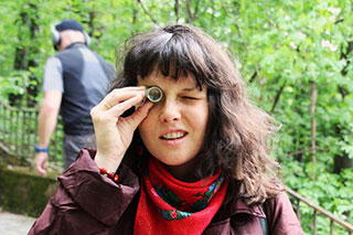 A person in a park with green trees, looking toward the camera through a small magnifying glass held to one eye.