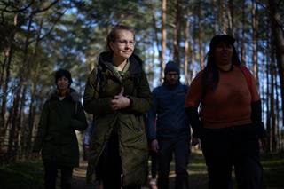 A group of people walking in the forest. Others are in the shadows, while a forest therapy guide is in the sunlight, focused and smiling.