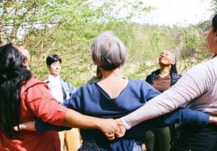 A group of people from different continents and with diverse skin tones standing in a forest, holding hands behind their backs in a circle, with relaxed faces and closed eyes.