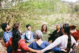 People from different continents holding hands in a circle with their eyes closed, standing in a forest.