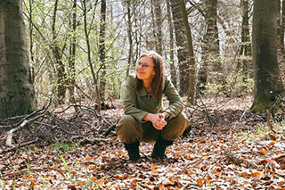 A forest therapy guide crouching in the forest, dressed in earthy colors, smiling.