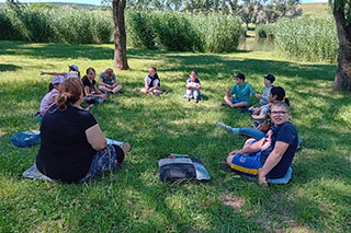On a green meadow among trees by a lake, elementary school students sit in a circle, wearing shirts and shorts. A forest therapy guide sits on the ground among them, everyone focused.