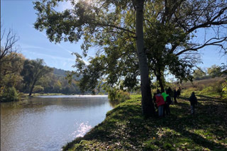 A landscape with a sunlit river, a tree on the riverbank, and in the distance, a group of people walking along the water.