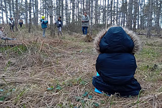 In a winter forest with leafless trees, adults and children are visible in the distance. In the foreground, a child in a winter hooded jacket sits on brown grass, seen from behind.