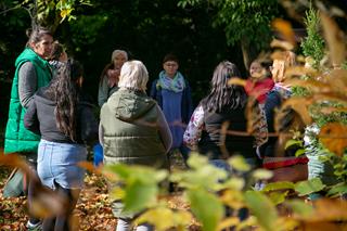 A group of women in winter jackets and coats standing in a circle, seen through grass in the shadows of forest trees.