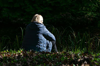 A woman in a winter jacket sitting on the ground with her back to us, among grass at the edge of a forest, illuminated by sunlight.