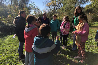 Children standing in a circle with their caregivers, dressed warmly in jackets, showing something to each other in the sunlight.