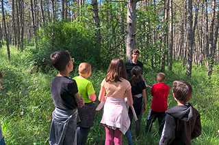 Children seen from behind in a green forest, looking at a person leading an exercise.