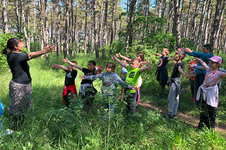 In green grass in a forest, a group of children imitates a person leading them, with arms stretched forward.