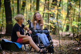 Two people in an autumn forest. One is sitting in a wheelchair, and the other is beside them.