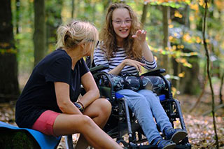 Two women in an autumn forest. One is sitting in a wheelchair, smiling and pointing something out to the other person sitting beside her.