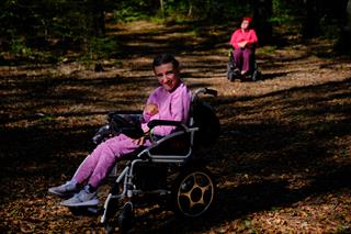 A forest path with a person in a wheelchair smiling at the camera, while another person in a wheelchair rests in the distance.