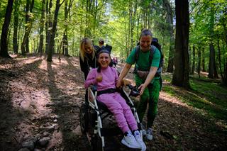 Descending a hill in the forest, a person in a wheelchair is assisted by two helpers, while more people walk in the distance