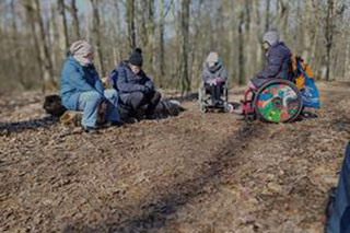 A winter forest with bare trees in the sunlight. Four people form a circle, dressed warmly in hats and jackets, with two of them in wheelchairs.