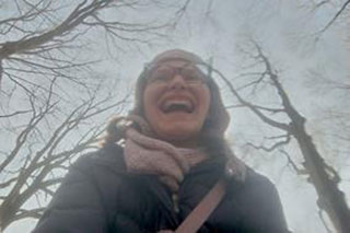 A person in a winter forest, seen from below in a close-up portrait, smiling widely, with tree branches and sky in the background