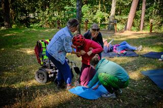 Sunny forest clearing with green trees. One person is lying on a mat, while two others assist a wheelchair user in moving onto a mat on the ground.
