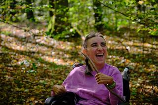 A person in a wheelchair in the forest, holding a stick and smiling while looking at someone off-frame.