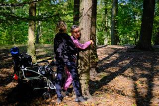 In a sunny forest, a person who previously sat in a wheelchair is now standing and hugging a tree, while another person assists them.
