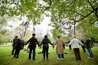 In a green park, under tree branches, older people stand in a large circle, wearing jackets and coats, holding hands, seen from behind