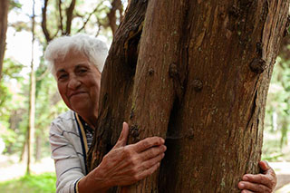 An older woman with gray hair hugging a large tree branch, her hands around the tree and smiling at the camera.