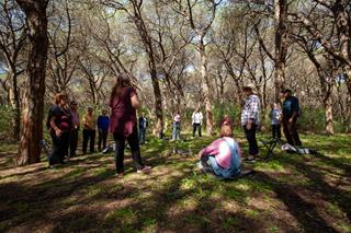 On a meadow among trees in a Mediterranean-like setting, a group of people stands in a line, one sits on the ground, listening to a person speaking.