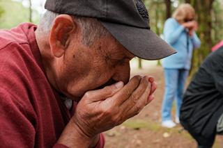 An older man wearing a cap smells something held in his hands behind his back, with other people also smelling objects in their hands.