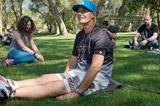 People sitting on green grass in various positions—legs stretched out, cross-legged, crouching—at different distances from each other, dressed for warm weather.