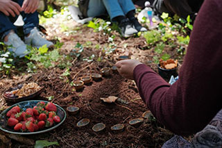 A picnic laid out on the ground: a bowl of strawberries and small clay cups arranged in a circle. A person's hand and feet are visible, as if people are sitting around.