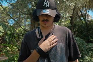 A man standing with a forest in the background, wearing a cap that shades his eyes, with his right hand on his chest.